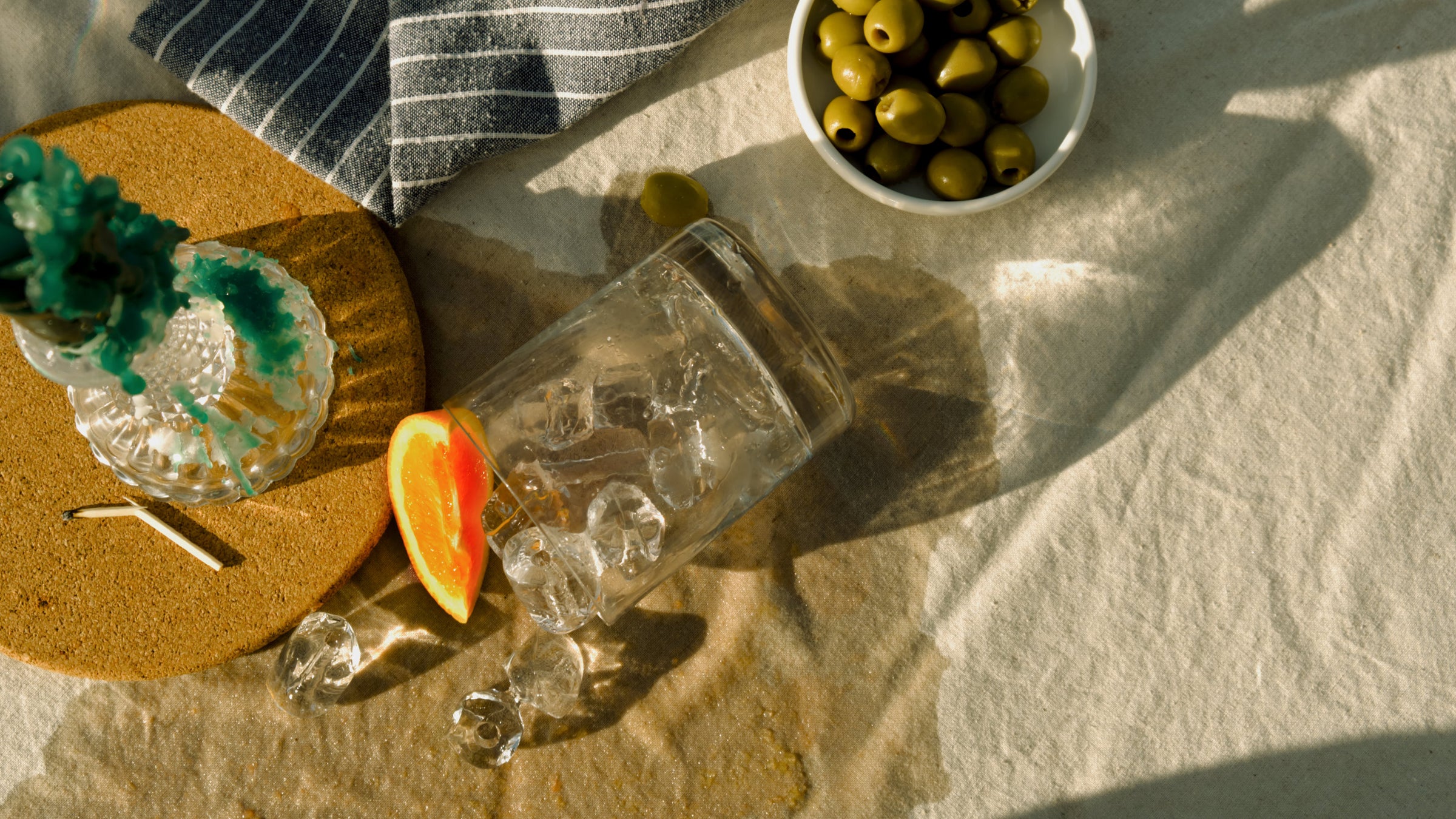 A spilled glass with ice and an orange slice, surrounded by green olives and a candle, creating a relaxed and natural atmosphere linked to Good Natured Spirits and Devon Gin.