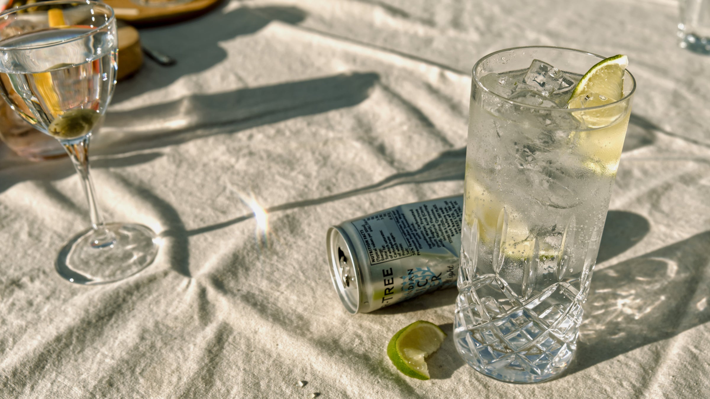 A tall glass of gin and tonic with ice and lime, set on a sunlit table with tonic water can in the background, showcasing a crisp and refreshing moment with Good Natured Spirits.