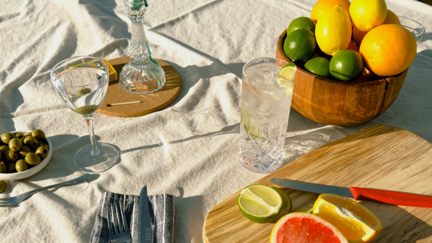 A close-up view of a sophisticated table spread of cocktails featuring Good Natured Spirits Devon gin cocktail, and a stylish wooden fruit bowl filled with citrus fruits.