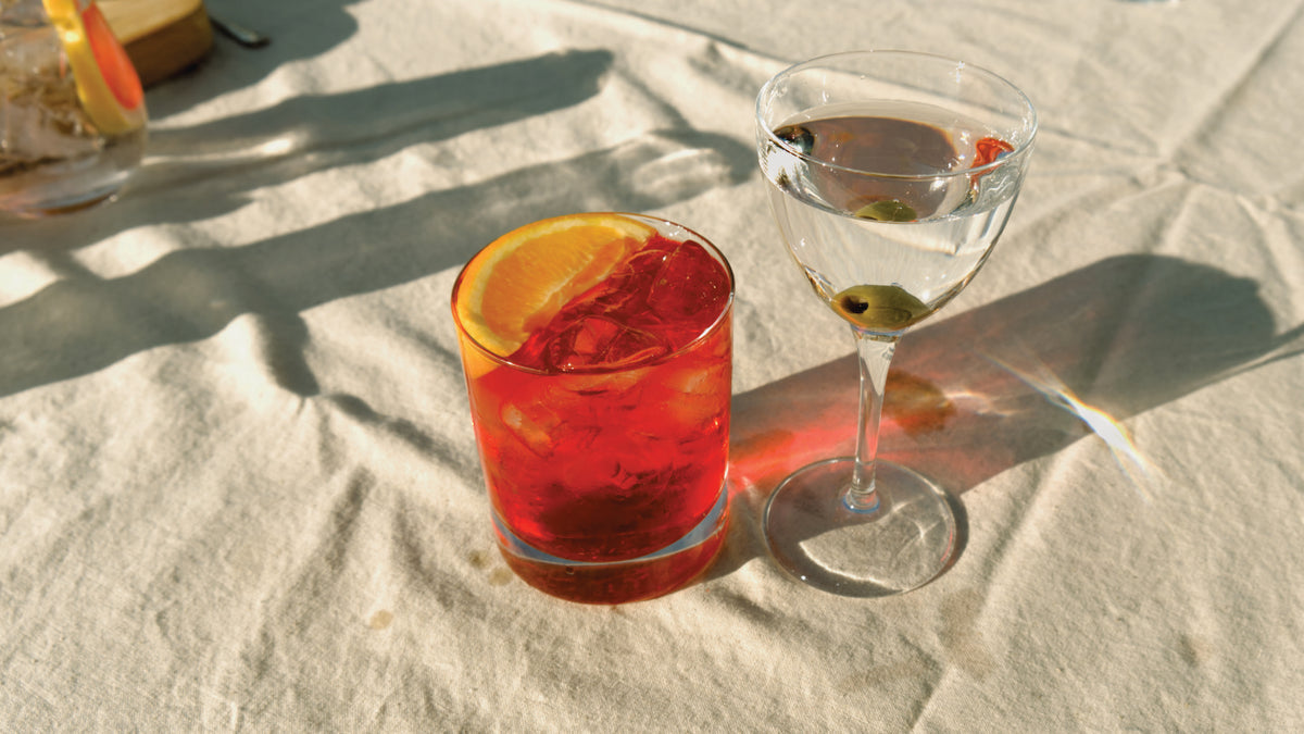 A vibrant and enticing negroni cocktail made with Good Natured Spirits Devon gin, accompanied by a martini made with Copper Frog Gin with olives on a white linen tablecloth. 