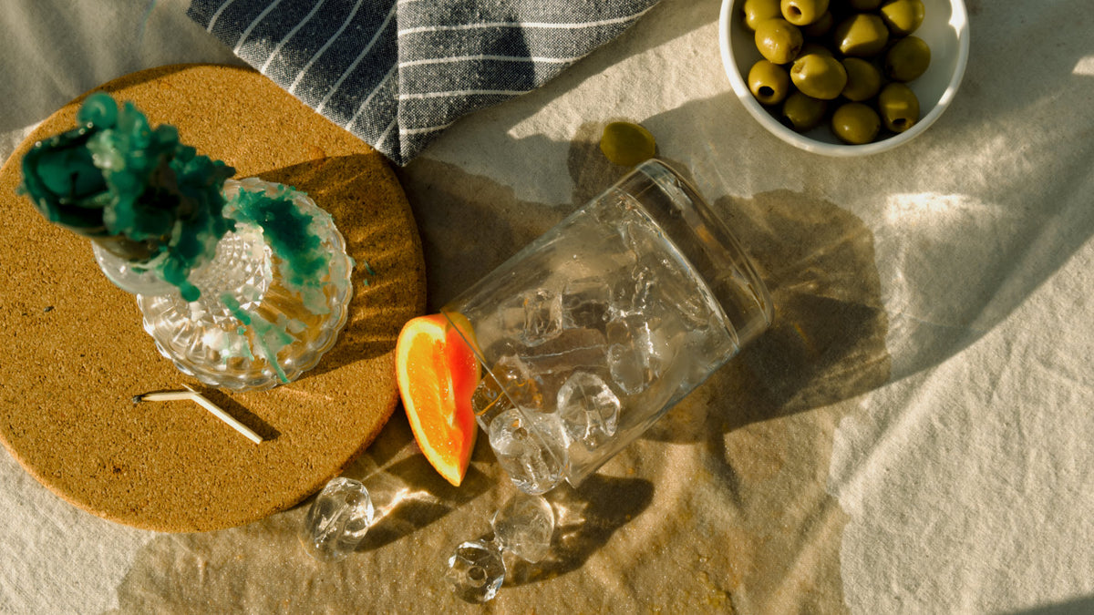 A spilled glass with ice and an orange slice, surrounded by green olives and a candle, creating a relaxed and natural atmosphere linked to Good Natured Spirits and Devon Gin.